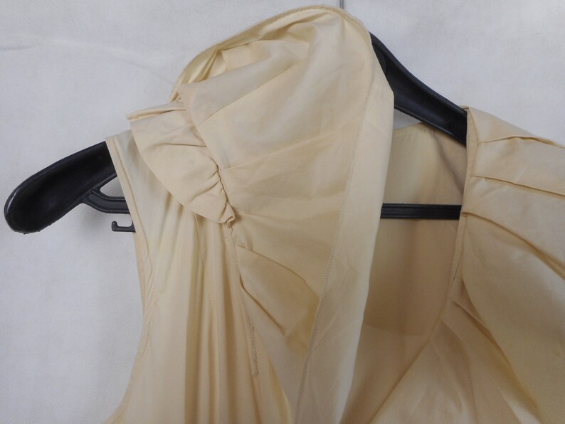 May include: A sleeveless, cream-colored blouse with a ruffled neckline and a draped design.