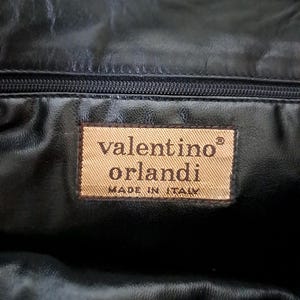 May include: A black leather interior lining featuring a rectangular tan fabric label with the text valentino orlandi and MADE IN ITALY printed in dark brown, positioned directly below a horizontal black zipper closure.