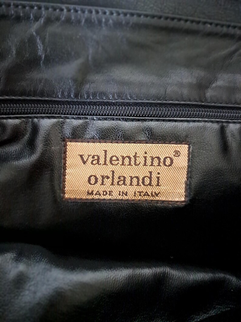 May include: A black leather interior lining featuring a rectangular tan fabric label with the text valentino orlandi and MADE IN ITALY printed in dark brown, positioned directly below a horizontal black zipper closure.