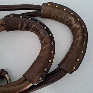 May include: Two brown leather handbag handles featuring a curved, padded design with visible wear and texture. Each handle is accented with a row of small, round, metallic brass-coloured studs along the outer edge, securing the leather overlay.