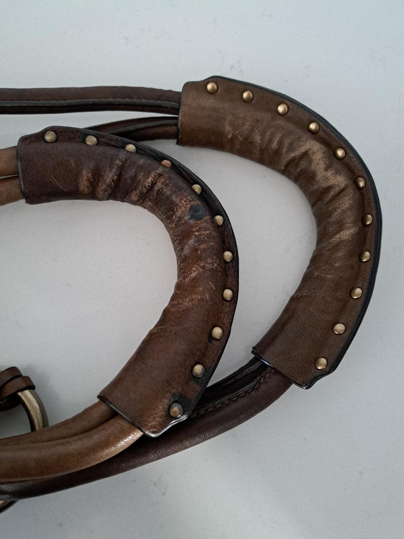 May include: Two brown leather handbag handles featuring a curved, padded design with visible wear and texture. Each handle is accented with a row of small, round, metallic brass-coloured studs along the outer edge, securing the leather overlay.