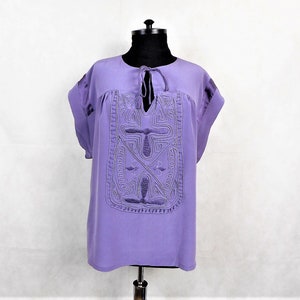 May include: A lavender blouse with a geometric design embroidered on the front. The blouse has a tie at the neckline and short sleeves.
