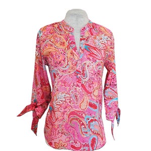 Könnte beinhalten: Eine rosa Paisley-Bluse mit langen Ärmeln und Schleifen an den Manschetten. Die Bluse hat einen Stehkragen und einen V-Ausschnitt. Das Paisley-Muster umfasst Rosa-, Rot-, Orange- und Blautöne.