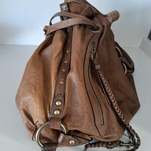 May include: A slouchy brown leather hobo bag featuring a braided leather shoulder strap, antique brass-toned metal hardware, a vertical zippered side pocket, and a decorative leather strip accented with a row of small round metal studs.