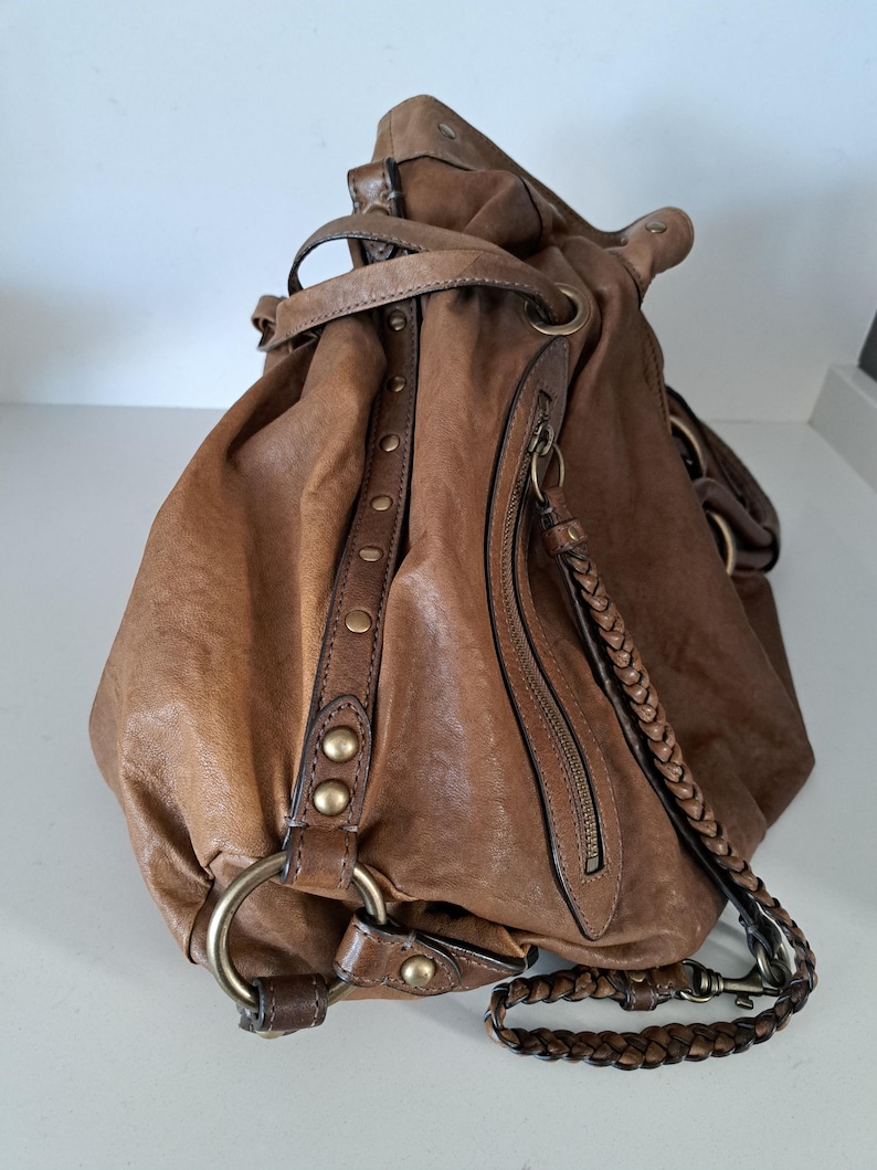 May include: A slouchy brown leather hobo bag featuring a braided leather shoulder strap, antique brass-toned metal hardware, a vertical zippered side pocket, and a decorative leather strip accented with a row of small round metal studs.