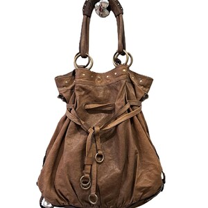 May include: A brown slouchy hobo bag featuring a gathered top, a drawstring closure with leather ties, and metallic ring hardware. The bag includes two structured handles with decorative studs and a textured finish throughout the exterior surface.