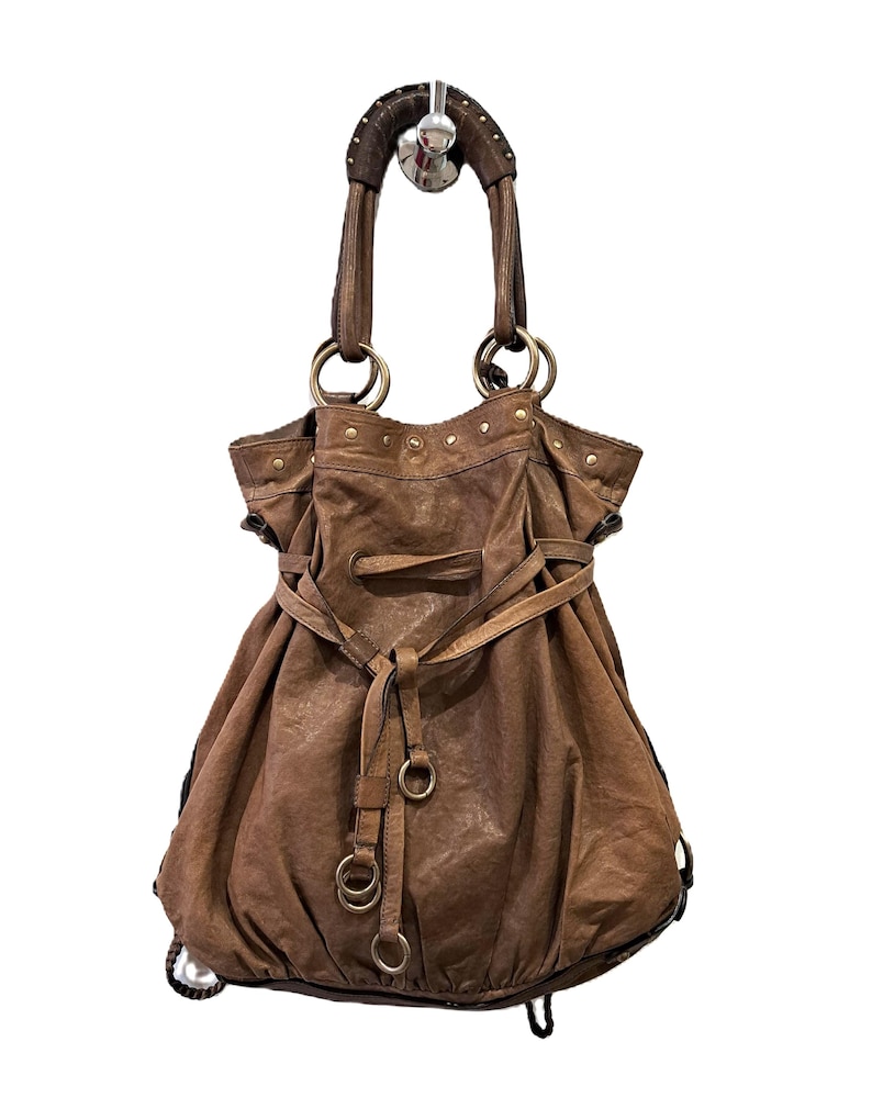 May include: A brown slouchy hobo bag featuring a gathered top, a drawstring closure with leather ties, and metallic ring hardware. The bag includes two structured handles with decorative studs and a textured finish throughout the exterior surface.