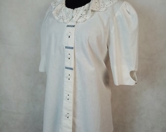 Pode incluir: Uma camisa branca de botões com colarinho de renda e acabamento azul e branco na frente. A camisa tem mangas curtas e está em um manequim.