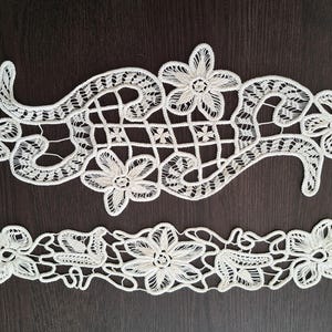 Op de afbeelding: Twee stukken witte kant met bloemenmotieven. De kant is delicaat en ingewikkeld, met een herhalend patroon van bloemen en bladeren.