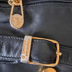 May include: A black leather handbag featuring gold-tone hardware. The zipper pull is engraved with the Valentino Orlandi logo. A rectangular metal buckle on a leather strap is also engraved with the text Valentino Orlandi and a registered trademark symbol.