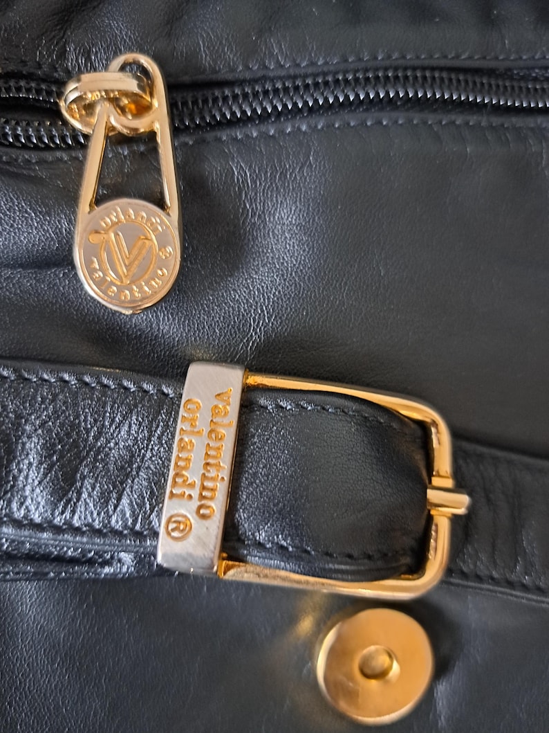 May include: A black leather handbag featuring gold-tone hardware. The zipper pull is engraved with the Valentino Orlandi logo. A rectangular metal buckle on a leather strap is also engraved with the text Valentino Orlandi and a registered trademark symbol.