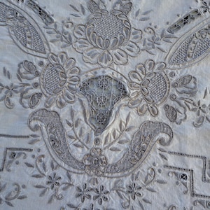 May include: A close-up of a white linen tablecloth with intricate gray embroidery. The embroidery features a floral design with delicate leaves and flowers.