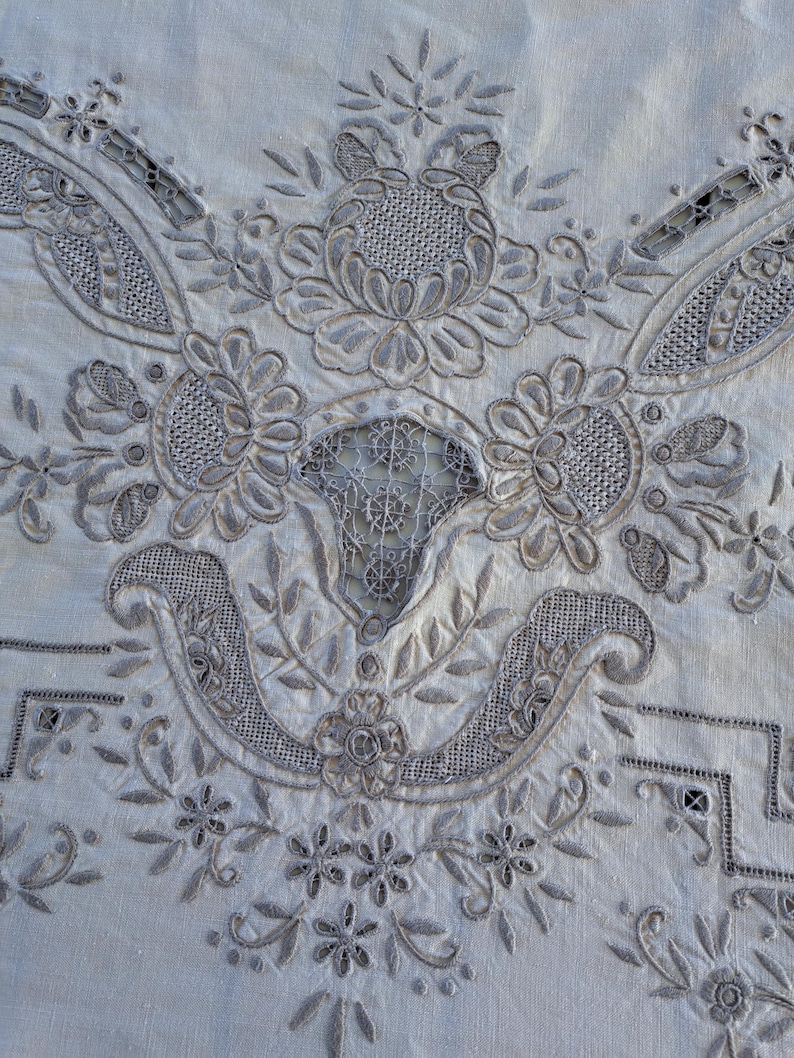 May include: A close-up of a white linen tablecloth with intricate gray embroidery. The embroidery features a floral design with delicate leaves and flowers.