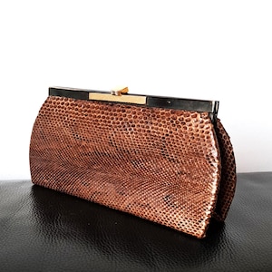 May include: A vintage brown clutch bag with a textured, reptile-like pattern. The rectangular bag has a black frame and a gold-coloured clasp. The clutch is set against a white background and rests on a black surface.