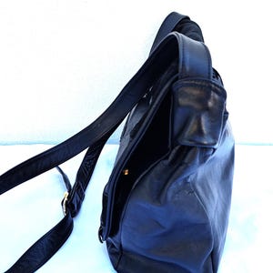 May include: A black leather shoulder bag with a smooth texture, featuring a long adjustable strap and a fold-over flap closure. The bag has a structured side profile with visible stitching and a small gold-toned metal hardware detail on the strap.