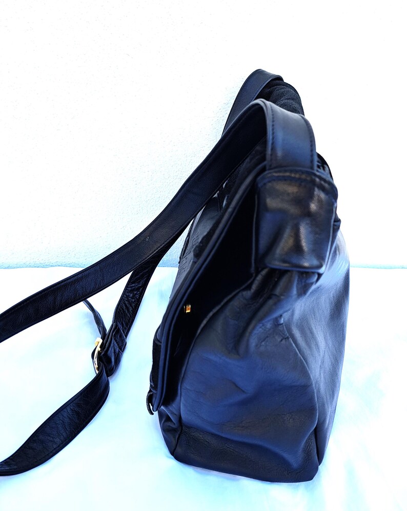 May include: A black leather shoulder bag with a smooth texture, featuring a long adjustable strap and a fold-over flap closure. The bag has a structured side profile with visible stitching and a small gold-toned metal hardware detail on the strap.