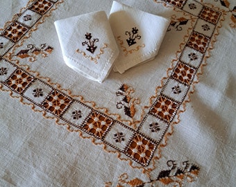 Può includere: Due tovaglioli di lino bianco con ricamo marrone di un motivo floreale, posizionati su una tovaglia bianca con un bordo ricamato marrone e arancione.