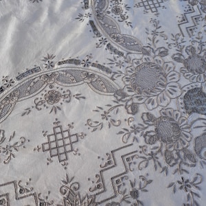 May include: A white tablecloth with intricate embroidery in a silver gray color. The embroidery features floral designs, geometric patterns, and a repeating motif of a square with a cross inside.