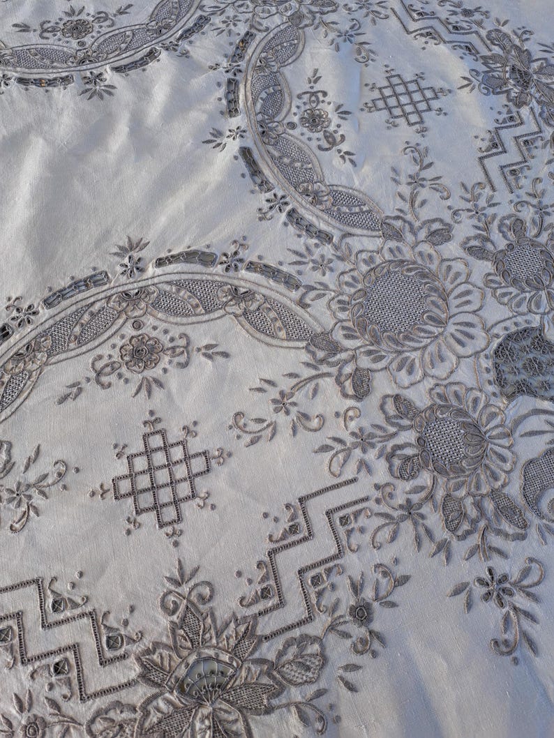 May include: A white tablecloth with intricate embroidery in a silver gray color. The embroidery features floral designs, geometric patterns, and a repeating motif of a square with a cross inside.