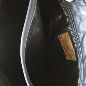 May include: The interior of a black bag featuring a central divider and a zippered pocket on one side. A rectangular brown leather patch is stitched onto the interior lining, displaying embossed text that is partially visible and worn.
