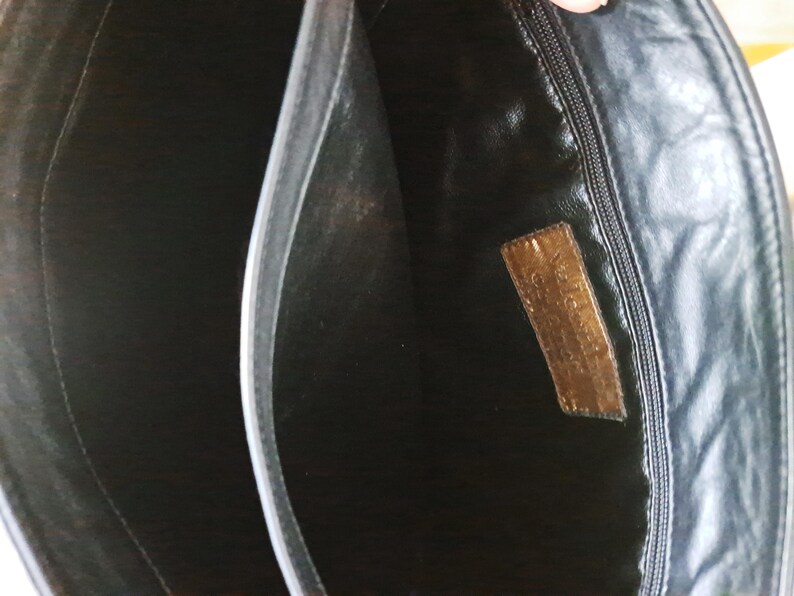 May include: The interior of a black bag featuring a central divider and a zippered pocket on one side. A rectangular brown leather patch is stitched onto the interior lining, displaying embossed text that is partially visible and worn.