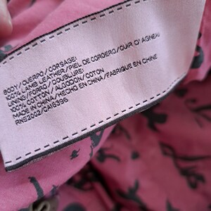 May include: A light pink fabric clothing label with black text stitched onto a pink lining featuring a black pattern. The label reads: BODY/CUERPO/CORSAGE: 100% LAMB LEATHER/PIEL DE CORDERO/CUIR D'AGNEAU, LINING/FORRO/DOUBLURE: 100% COTTON/ALGODON/COTON, MADE IN CHINA/HECHO EN CHINA/FABRIQUE EN CHINE, RNS2002/CA16396.