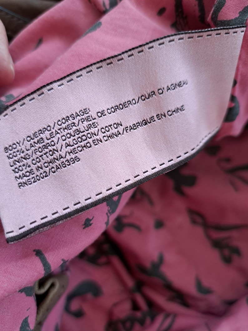 May include: A light pink fabric clothing label with black text stitched onto a pink lining featuring a black pattern. The label reads: BODY/CUERPO/CORSAGE: 100% LAMB LEATHER/PIEL DE CORDERO/CUIR D'AGNEAU, LINING/FORRO/DOUBLURE: 100% COTTON/ALGODON/COTON, MADE IN CHINA/HECHO EN CHINA/FABRIQUE EN CHINE, RNS2002/CA16396.
