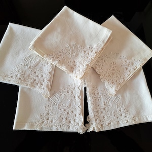 May include: Four white linen napkins with detailed lace edging and embroidered patterns. The napkins are folded and arranged, highlighting the delicate craftsmanship and classic design. The napkins are a soft white colour.