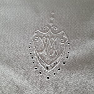 May include: Close-up of a white textile with an embroidered crest. The crest features an ornate design with the letters "WM" in the center, surrounded by decorative swirls and small, evenly spaced holes. The fabric has a subtle herringbone pattern.