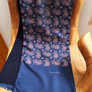 May include: A blue silk scarf with a paisley pattern and a Christian Dior logo. The scarf has a fringed edge.