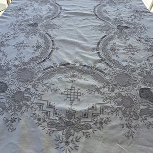 May include: A white tablecloth with intricate embroidered floral patterns in a gray color. The tablecloth has a delicate, lacy design and a slightly textured surface.