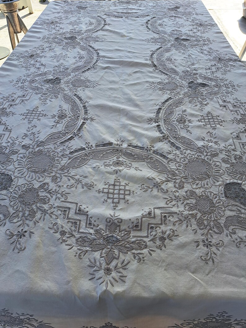May include: A white tablecloth with intricate embroidered floral patterns in a gray color. The tablecloth has a delicate, lacy design and a slightly textured surface.