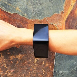 Può includere: Un ampio bracciale rigido nero a tinta unita con una finitura liscia e molto lucida. Il bracciale presenta una forma rettangolare e architettonica che avvolge il polso, mostrando una superficie riflettente che cattura la luce sul suo esterno scuro.