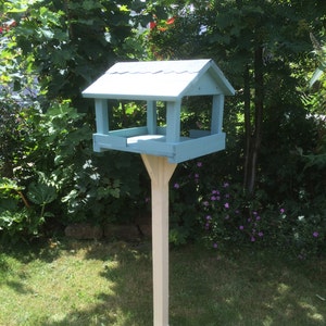 May include: A light blue bird feeder with a white roof and a white wooden stand with a cross-shaped base. The feeder is in a grassy area with green foliage in the background.