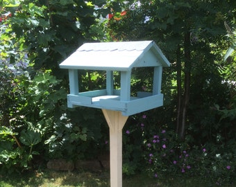 May include: A light blue bird feeder with a white roof and a white wooden stand with a cross-shaped base. The feeder is in a grassy area with green foliage in the background.