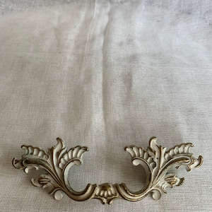 May include: Ornate, antique-style drawer pull with a white and gold finish. The handle features a curved design with detailed floral and leaf motifs. The metal hardware is likely made of brass or a similar material, adding a vintage aesthetic.