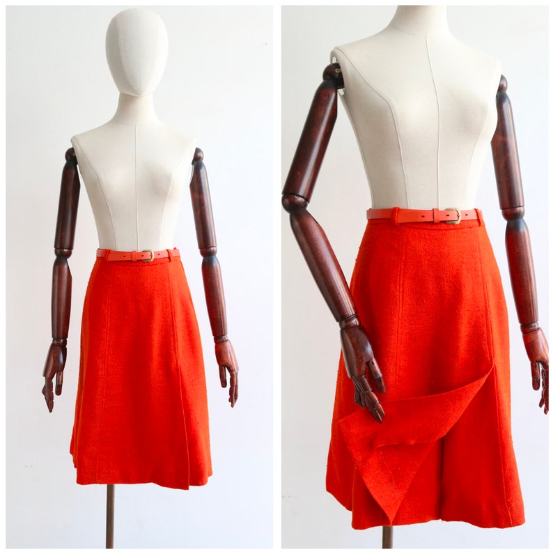 May include: A bright orange A-line skirt with a brown leather belt. The skirt is made of a textured fabric and has a slight flare at the hem.