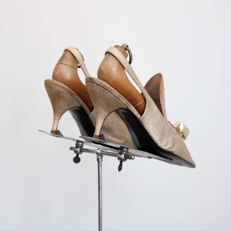 May include: A pair of gold slingback heels with pointed toes and small heels. The shoes are displayed on a metal stand with wooden shoe forms inside. The heels have a bow detail on the front.