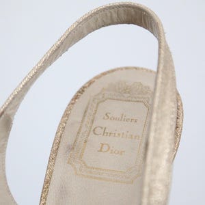 May include: Close-up of a gold-colored slingback shoe. The interior sole features the text "Souliers Christian Dior" in gold lettering within a decorative frame. The shoe's strap is visible, showcasing a textured design.