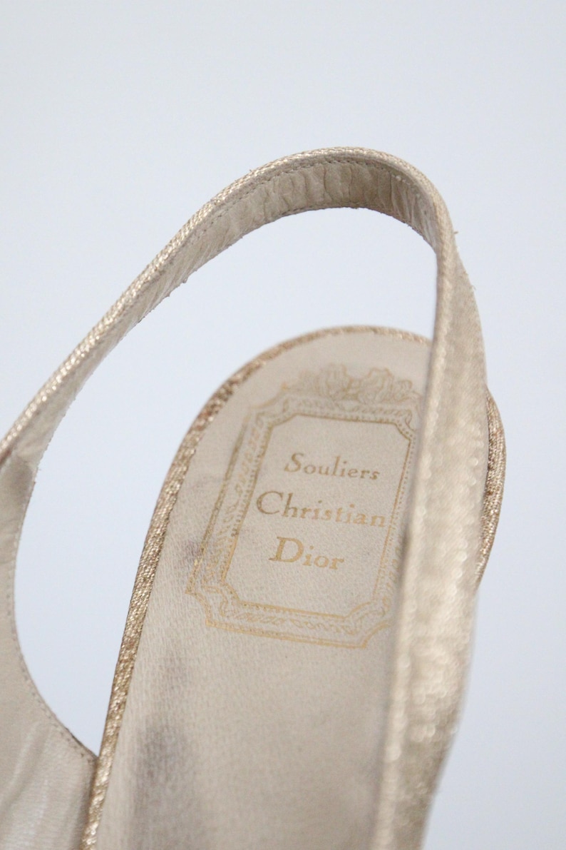 May include: Close-up of a gold-colored slingback shoe. The interior sole features the text "Souliers Christian Dior" in gold lettering within a decorative frame. The shoe's strap is visible, showcasing a textured design.