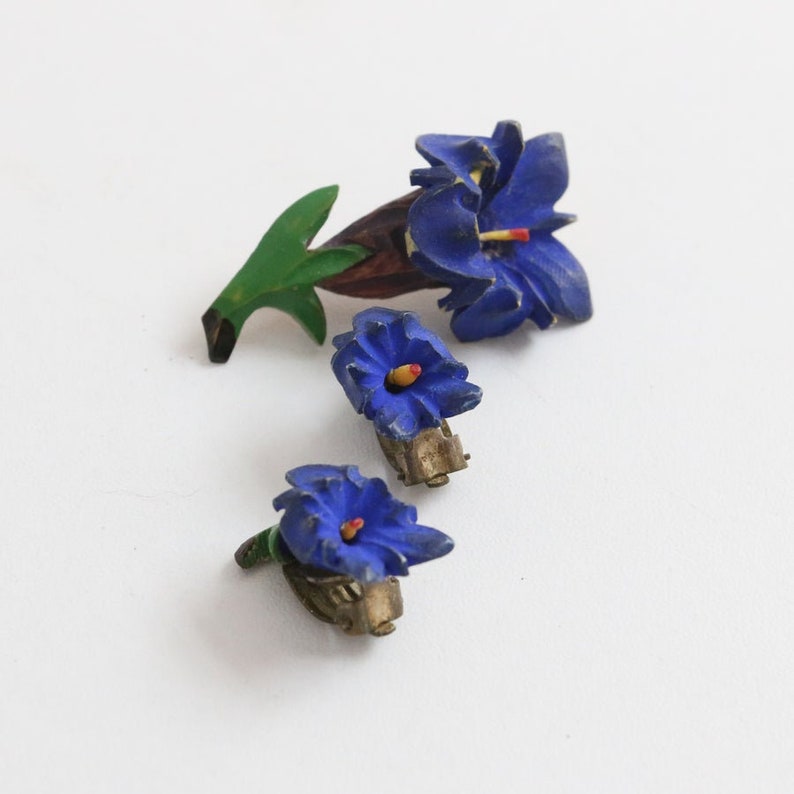 May include: A vintage jewelry set featuring a blue flower brooch and matching clip-on earrings. The brooch has a green leaf and brown stem. The earrings and brooch have yellow and red accents. The set is likely made of painted metal.
