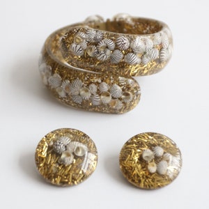 May include: A clear resin bracelet and matching earrings set. The jewelry is filled with gold glitter and small, white and black striped seashells. The bracelet is a spiral design, and the earrings are round.
