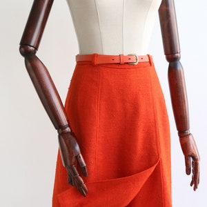 May include: A bright orange skirt with a fitted waist and a slightly flared hem. The skirt is cinched with a thin orange leather belt.