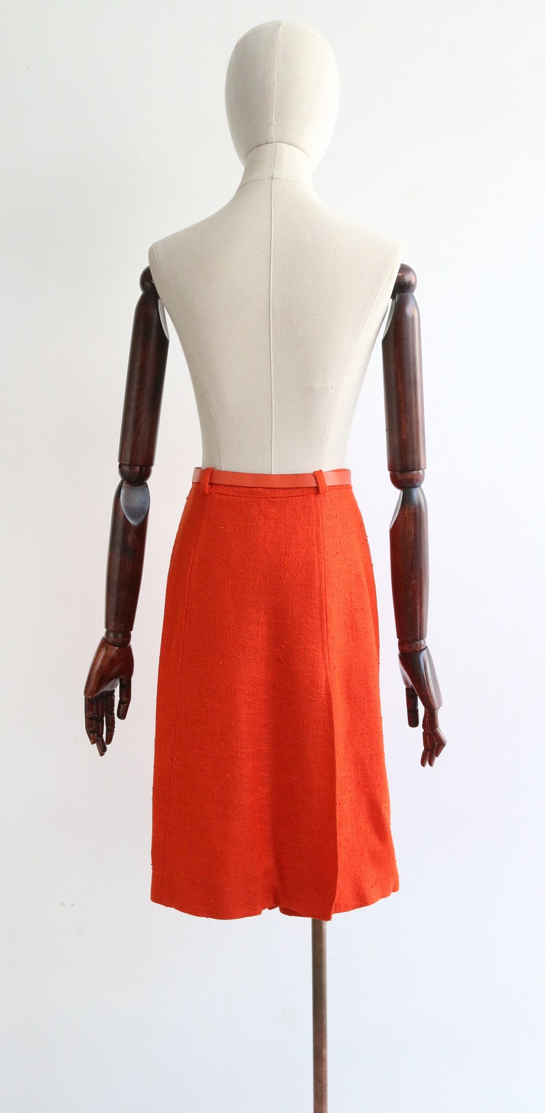 May include: A bright orange skirt with a red belt. The skirt is made of a textured fabric and has a straight silhouette.