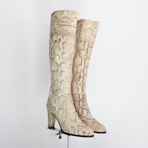 Puede incluir: Un par de botas altas hasta la rodilla con estampado de piel de serpiente en beige y crema. Las botas tienen una cremallera lateral y un tacón grueso. Se exhiben sobre un soporte metálico sobre un fondo blanco.