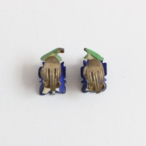 May include: A pair of vintage clip-on earrings. Each earring features a blue floral design with a green leaf accent and a gold-tone clip. The earrings are set against a white background.