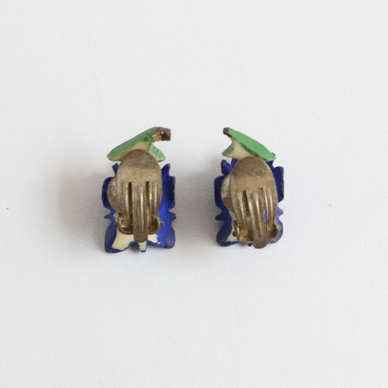 May include: A pair of vintage clip-on earrings. Each earring features a blue floral design with a green leaf accent and a gold-tone clip. The earrings are set against a white background.