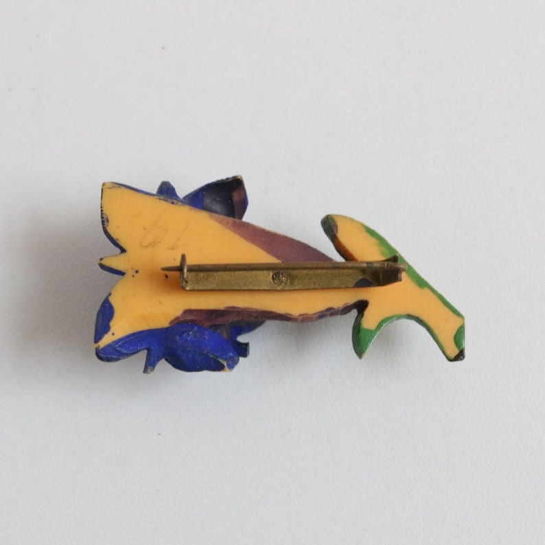 May include: A vintage brooch with a floral design. The brooch features a yellow base with blue, purple, and green accents. The pin is made of a metal alloy and has a simple clasp. The brooch is likely from the mid-20th century.