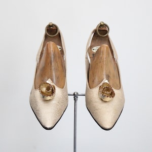 May include: A pair of gold-colored, pointed-toe pumps with a decorative floral embellishment on the vamp. The shoes are displayed on a metal stand, with wooden shoe forms inside. The heels are approximately 3 inches high.