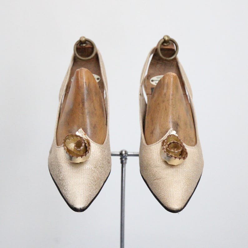 May include: A pair of gold-colored, pointed-toe pumps with a decorative floral embellishment on the vamp. The shoes are displayed on a metal stand, with wooden shoe forms inside. The heels are approximately 3 inches high.